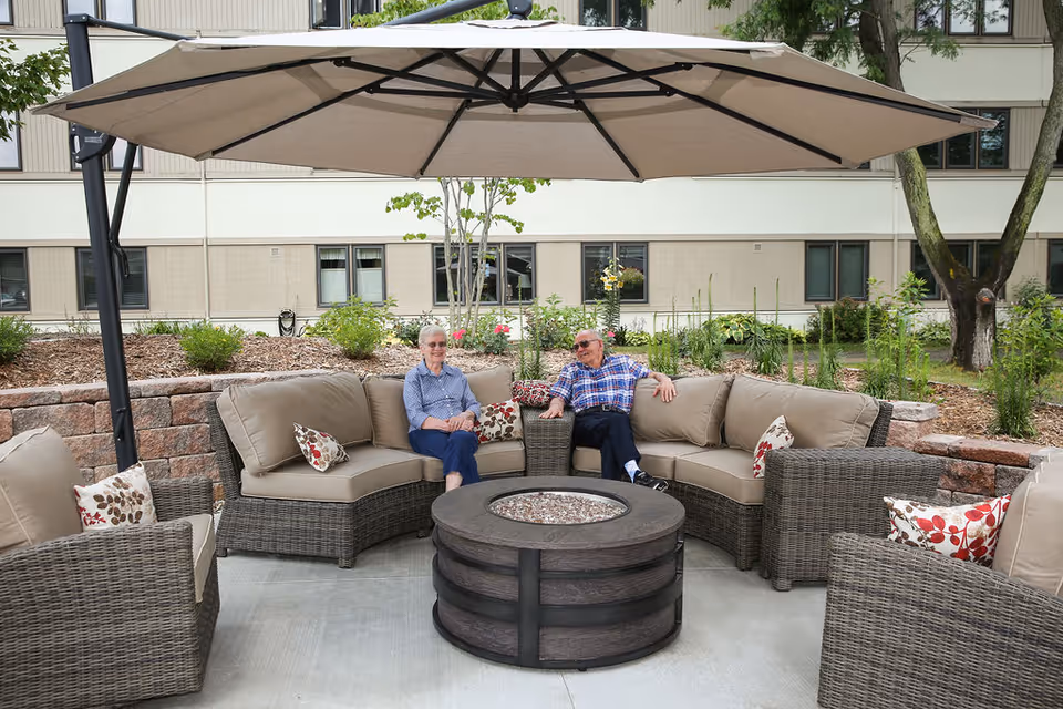 Two people seated on wicker patio furniture around a round fire table under a large umbrella in an outdoor courtyard in front of a multi-story building.