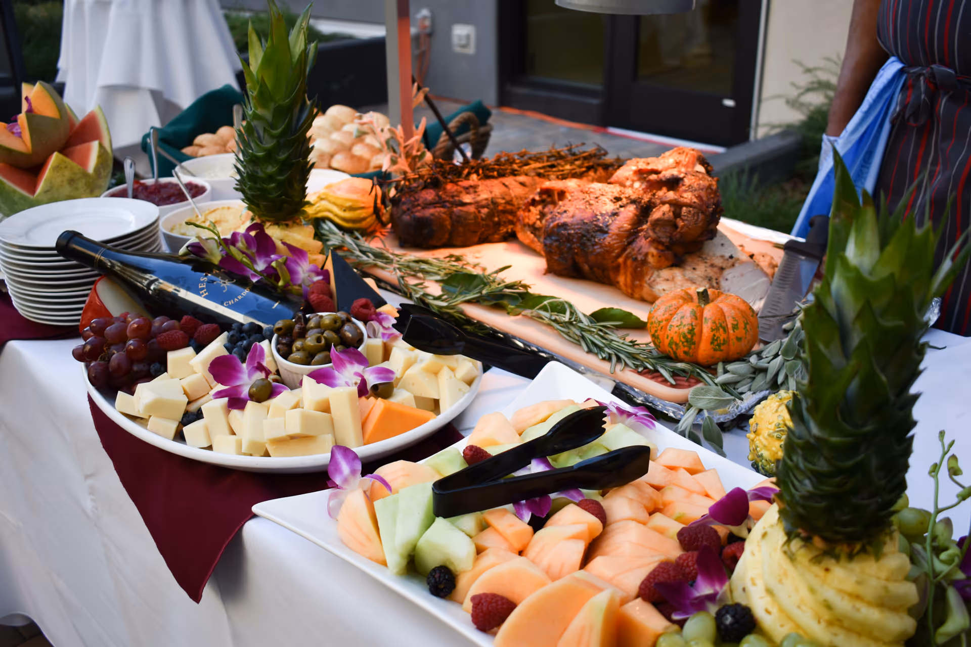 Outdoor buffet table with a carved roast, platters of fruit and cheese, and decorative pineapples.