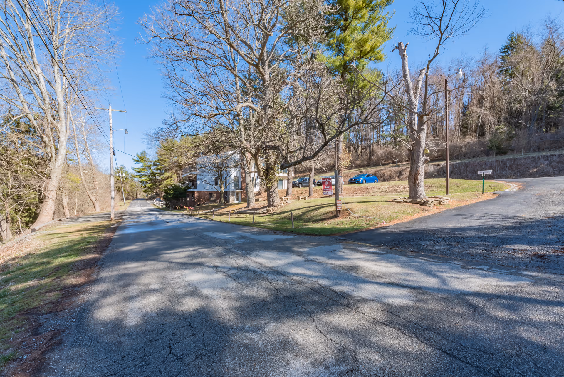 A paved road leading to a building partially visible on the left side, surrounded by leafless trees and some green grass patches under a clear blue sky. There are a few parked cars near the building and a signpost on the grassy area.