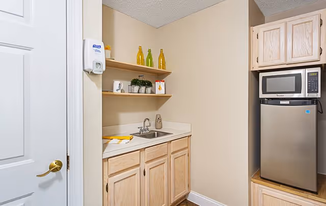 Small kitchenette area with light wood cabinets, a stainless steel sink, a countertop with a yellow and white towel, two wooden shelves holding decorative bottles and small plants, a microwave oven placed on top of a mini refrigerator, and a white door with a gold handle on the left side.
