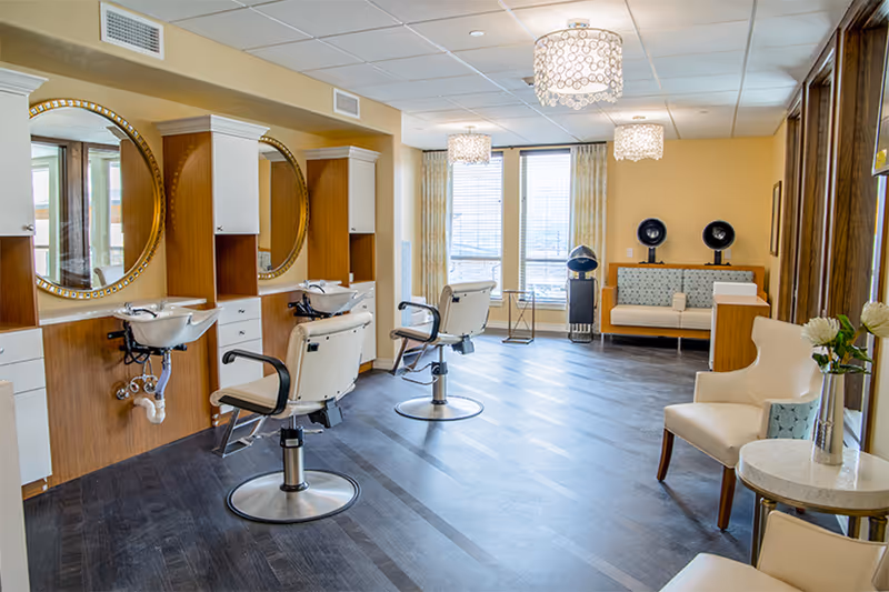 Interior hair salon with styling chairs, wash sinks, round mirrors, and a small waiting area in a senior living facility.