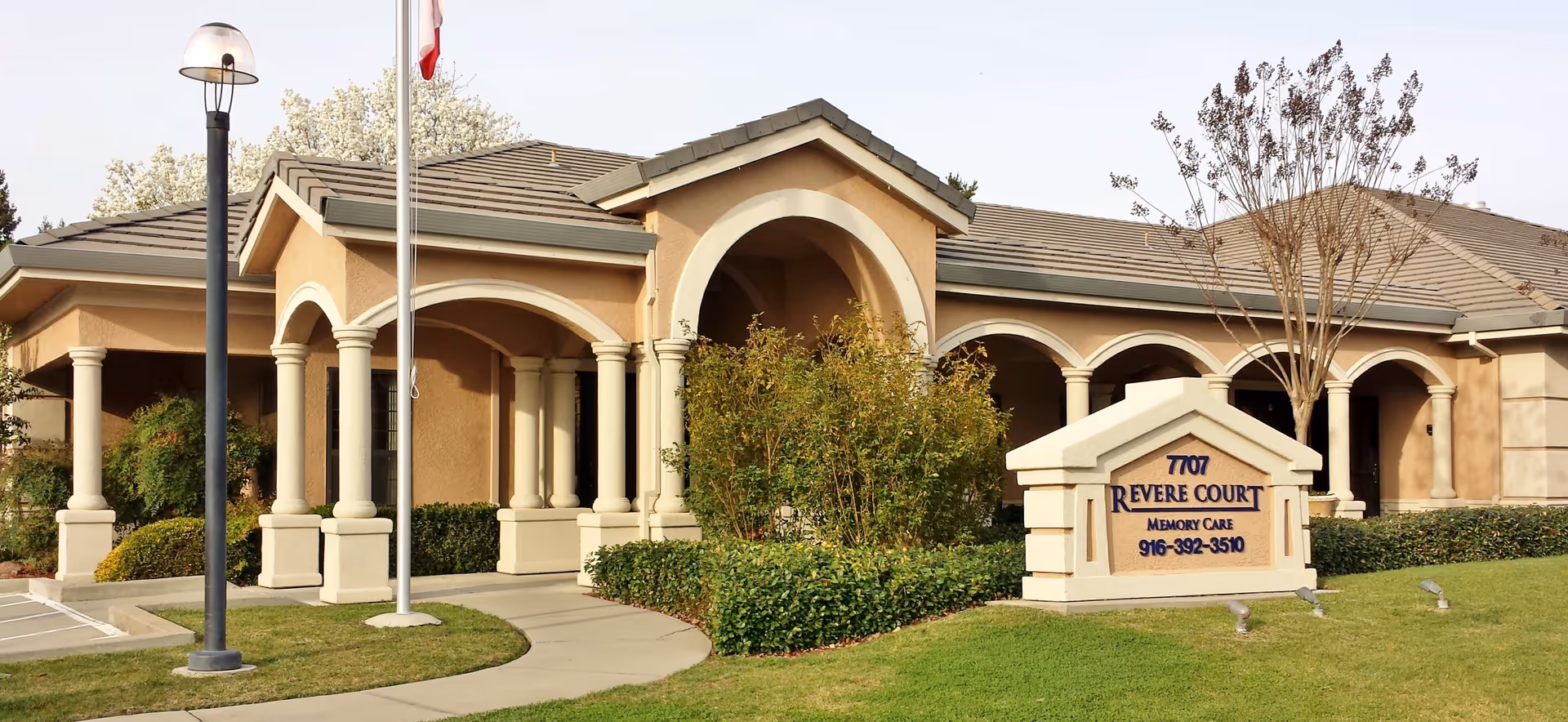Exterior view of Revere Court Memory Care facility showing a beige building with arched entrances and columns, surrounded by green bushes and a well-maintained lawn. A sign in front displays the facility name, address, and phone number. A flagpole and a streetlamp are also visible.