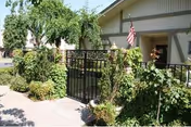 Entrance of a single-story building with an American flag, black metal fence, and lush greenery along a sidewalk.