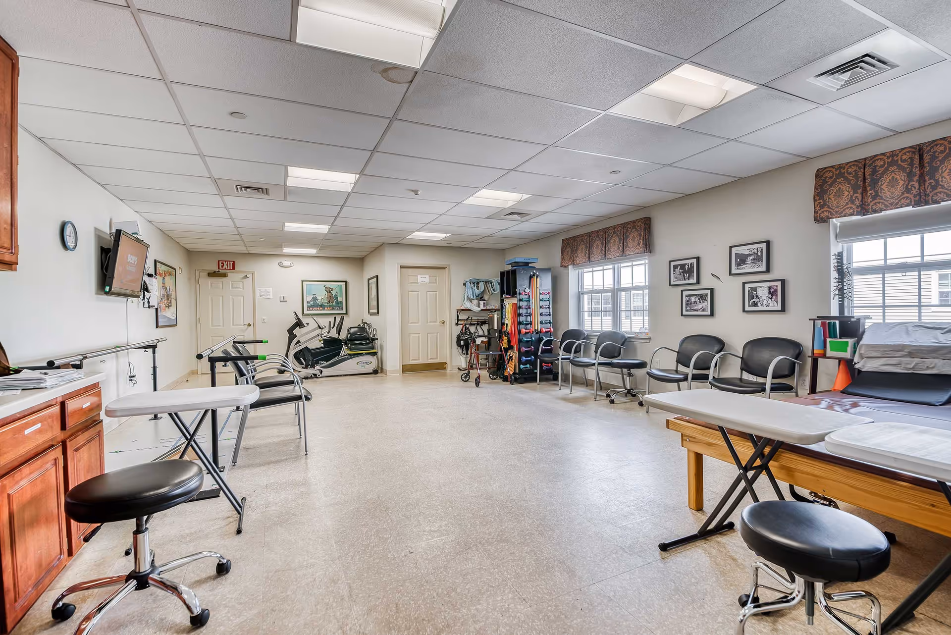 A spacious rehabilitation or therapy room with exercise equipment including stationary bikes, therapy tables, chairs along the walls, and various therapy tools and supplies. The room has a tiled floor, white ceiling with fluorescent lights, and windows with patterned valances letting in natural light.
