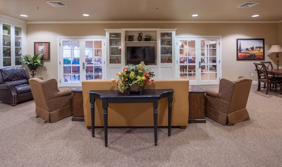 A cozy senior living common area with a tan sofa, two brown armchairs, and a dark wooden table with a floral arrangement on it. Behind the sofa is a white built-in entertainment center with a TV and shelves. The room has beige walls and carpet, with glass double doors leading to another room with tables and chairs. There is a leather armchair and a potted plant on the left side, and a dining table with chairs and a lamp on the right side.