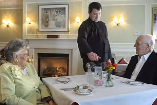 An elderly woman and man seated at a table in a dining room being served by a waiter in front of a fireplace.