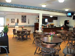 Interior view of a dining room with several round wooden tables and chairs. Each table has a small vase with flowers. The room has light-colored walls with decorative border trim near the ceiling, ceiling lights, and a television mounted in the corner. There is a counter area with a coffee machine and other items in the background.