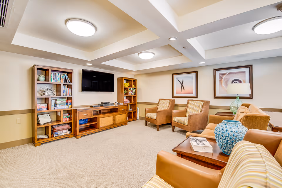 A cozy living room area with beige walls and carpet, featuring a flat-screen TV mounted on the wall between two wooden bookshelves filled with books and board games. There are four comfortable armchairs with striped cushions and a wooden coffee table with a blue decorative vase and a book. Two framed abstract artworks hang on the wall, and a table lamp with a glass base is placed on a side table next to a sofa.