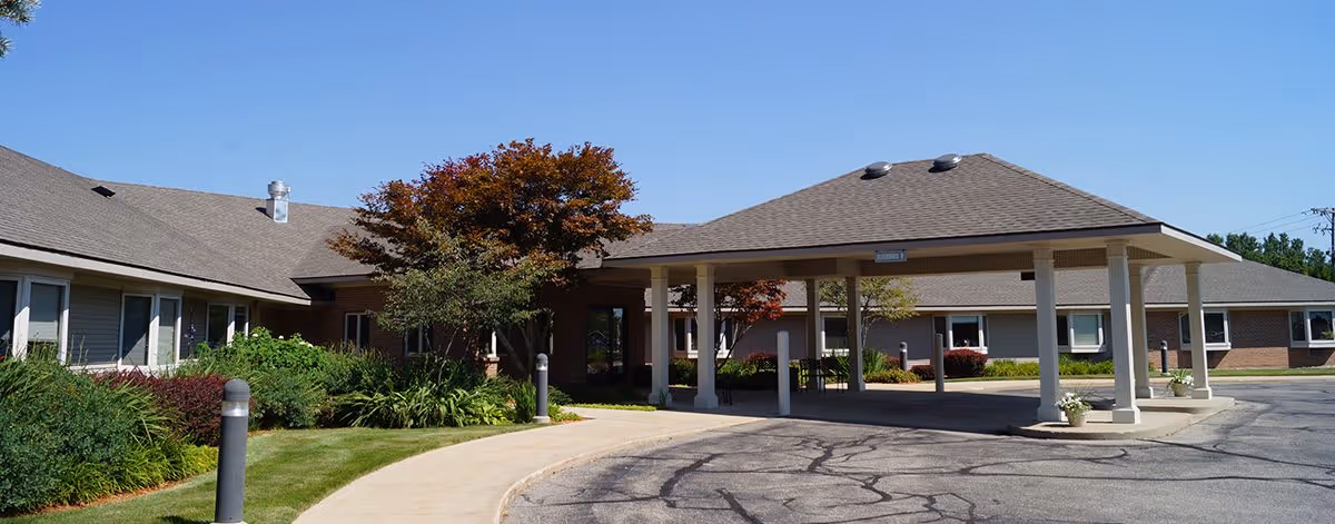 Exterior view of Railside Assisted Living Center showing a covered entrance with a driveway, surrounded by well-maintained landscaping including green bushes and trees under a clear blue sky.