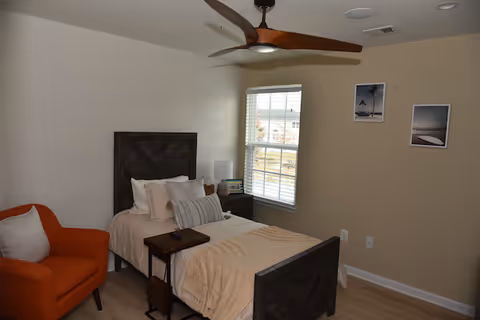 A bedroom with a single bed featuring a dark wooden headboard and footboard, beige bedding, and multiple pillows. Next to the bed is a small dark wooden nightstand with a lamp and decorative items. An orange armchair with a white pillow is positioned to the left of the bed. The room has light-colored walls, a window with white blinds, three framed pictures on the wall, and a ceiling fan with wooden blades.