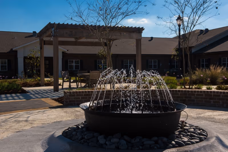 Outdoor courtyard area with a circular water fountain in the foreground surrounded by smooth stones. Behind the fountain is a wooden pergola with chairs and tables underneath. The building of the facility is visible in the background under a clear blue sky.