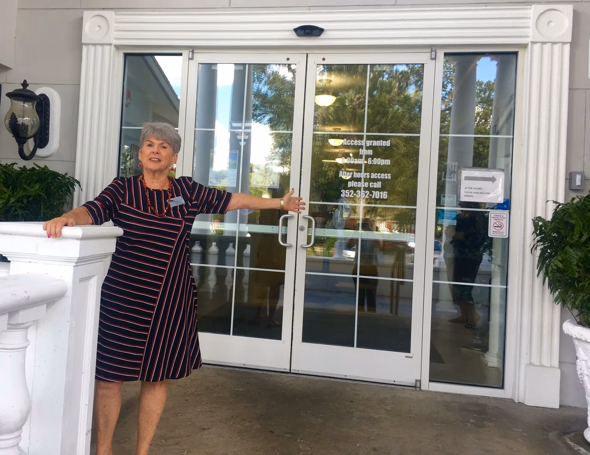 A woman with short gray hair wearing a black dress with red and white stripes stands outside a building entrance with double glass doors. She is gesturing with her right arm towards the doors. The entrance has white decorative columns and plants on either side. Text on the glass doors provides access hours and a phone number for after-hours access.