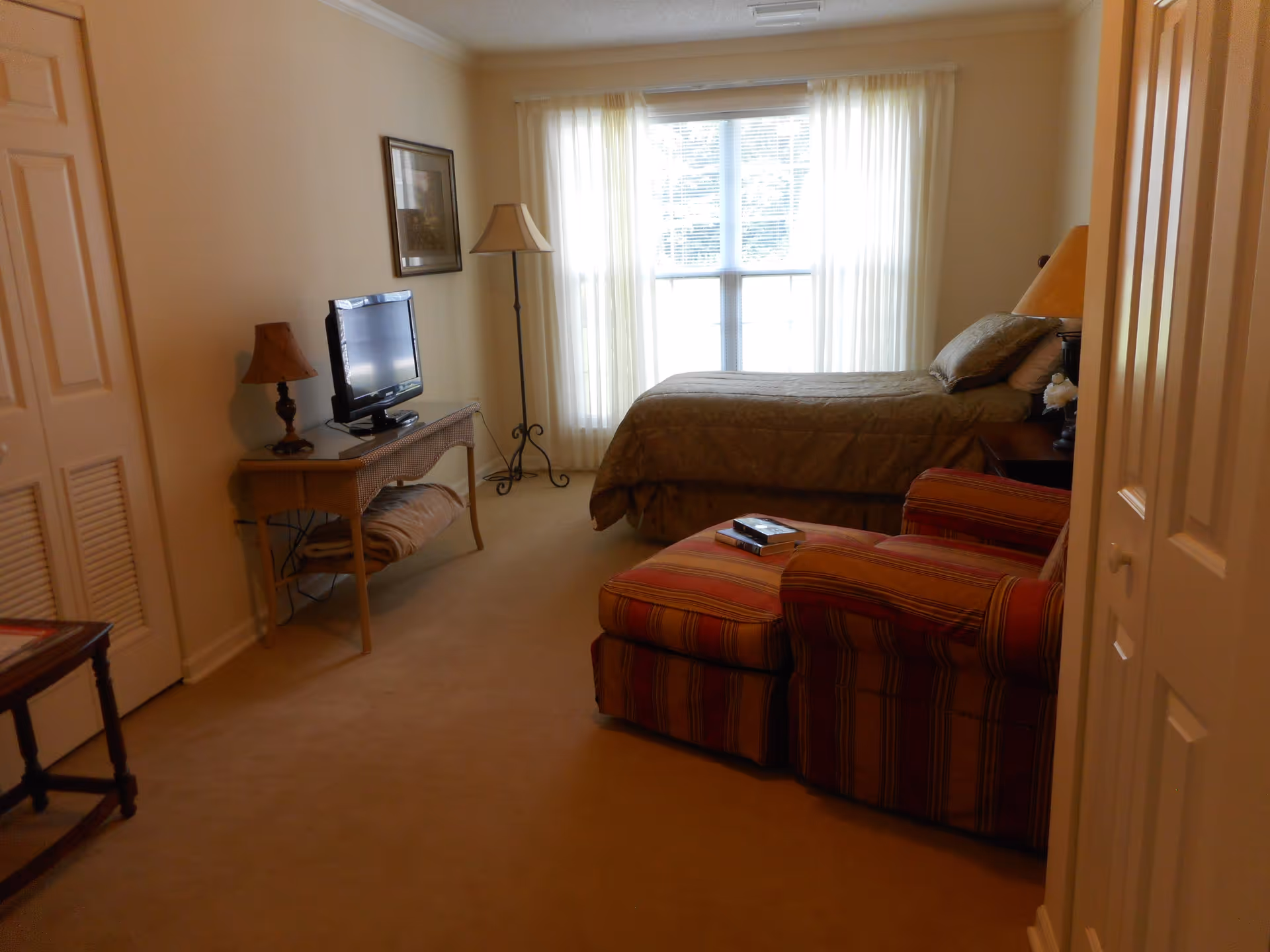 A cozy bedroom with a bed covered in a brown quilt, a striped armchair with an ottoman, a TV on a wicker console table, two lamps, a framed picture on the wall, and a large window with sheer white curtains letting in natural light.