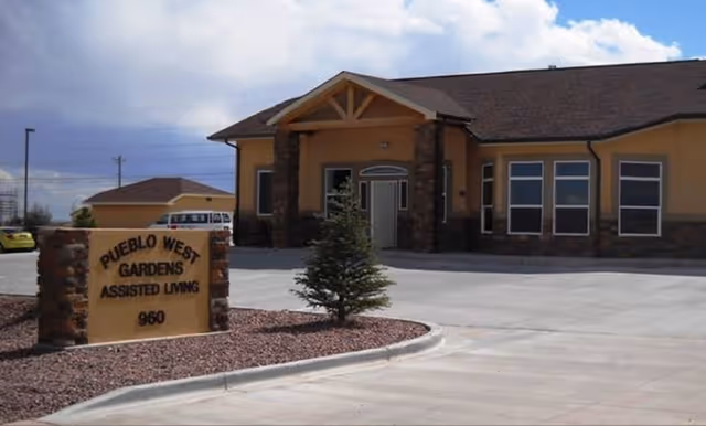 A stone entrance sign reading 'Pueblo West Gardens Assisted Living 960' sits by a paved driveway in front of a single-story assisted living building.