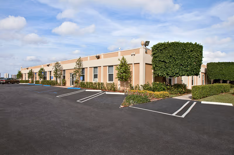 Single-story beige building with trimmed trees, landscaped beds and an empty parking lot under a partly cloudy sky.