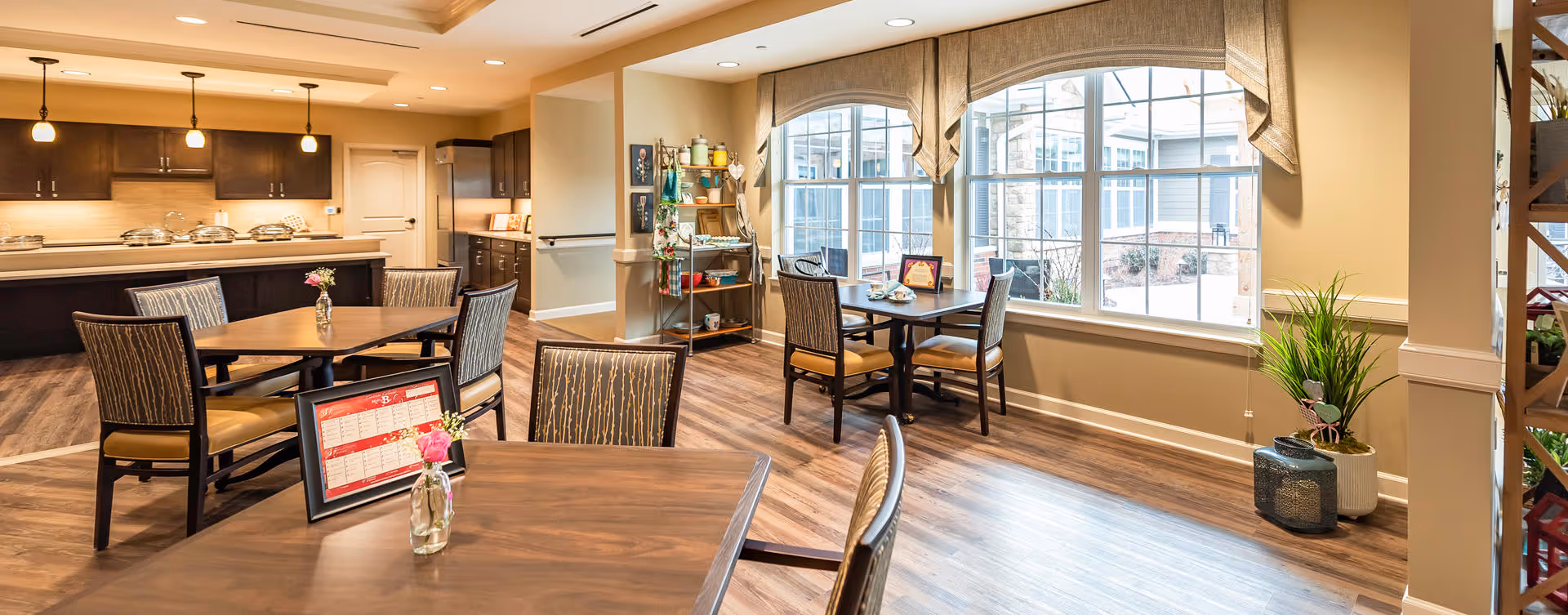 A bright and spacious dining area in a senior living facility with wooden tables and chairs. The room features large arched windows with beige valances allowing natural light to fill the space. There are small flower vases and framed menus on the tables. The background shows a kitchen area with dark wood cabinets and a counter with covered food trays. A small shelving unit with various items is positioned near the windows, and a potted plant sits on the floor by the window.