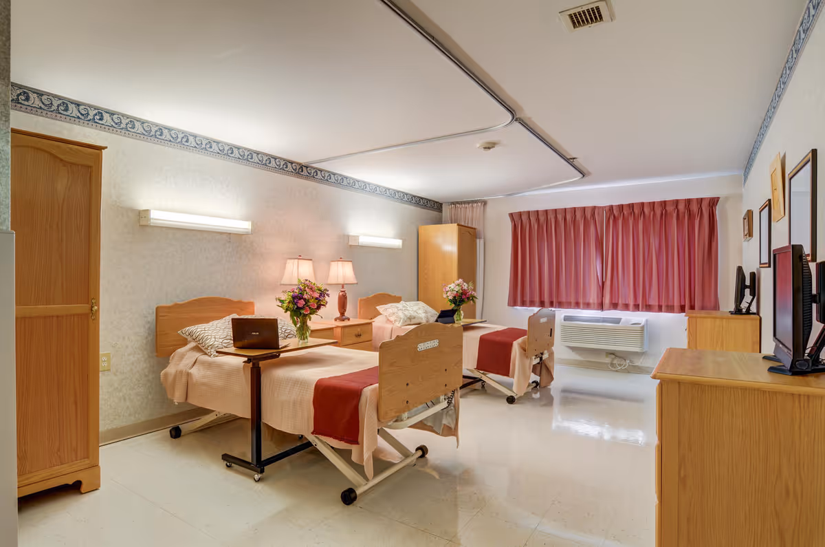 A well-lit nursing facility bedroom with two hospital-style beds, each with a red blanket and beige bedding. There are bedside tables with lamps and flower vases, a wooden wardrobe, a window with red curtains, and a TV on a wooden dresser. The room has a clean, simple design with light-colored walls and flooring.