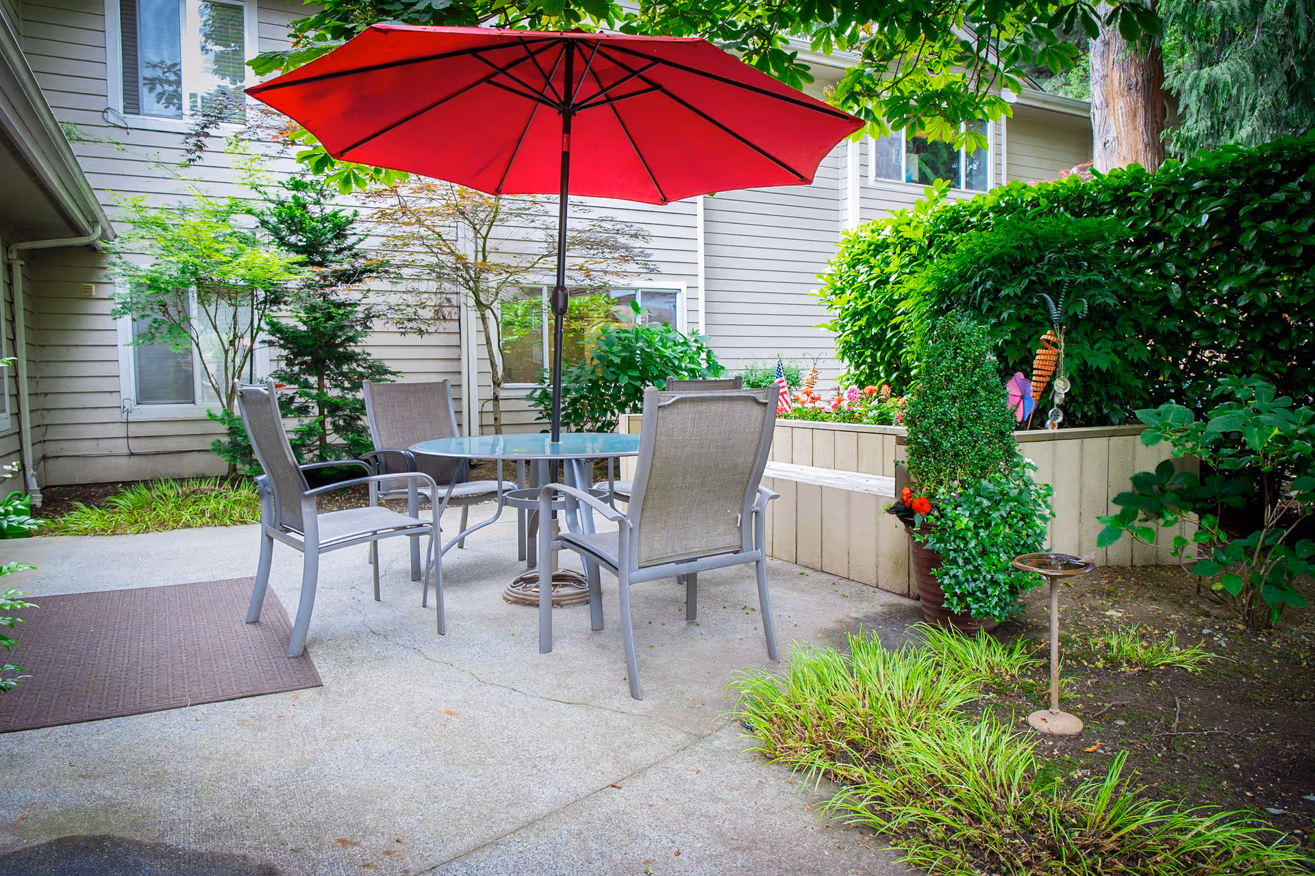 Outdoor patio area with a round glass table and four chairs under a large red umbrella. The patio is surrounded by greenery, including bushes, small trees, and plants. The exterior of a light-colored building is visible in the background.