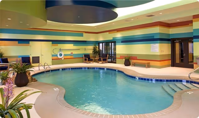 Indoor swimming pool with curved edges surrounded by a beige deck. The walls are decorated with horizontal stripes in various colors including green, blue, orange, and yellow. There are chairs and potted plants around the pool area, and doors leading to other rooms.