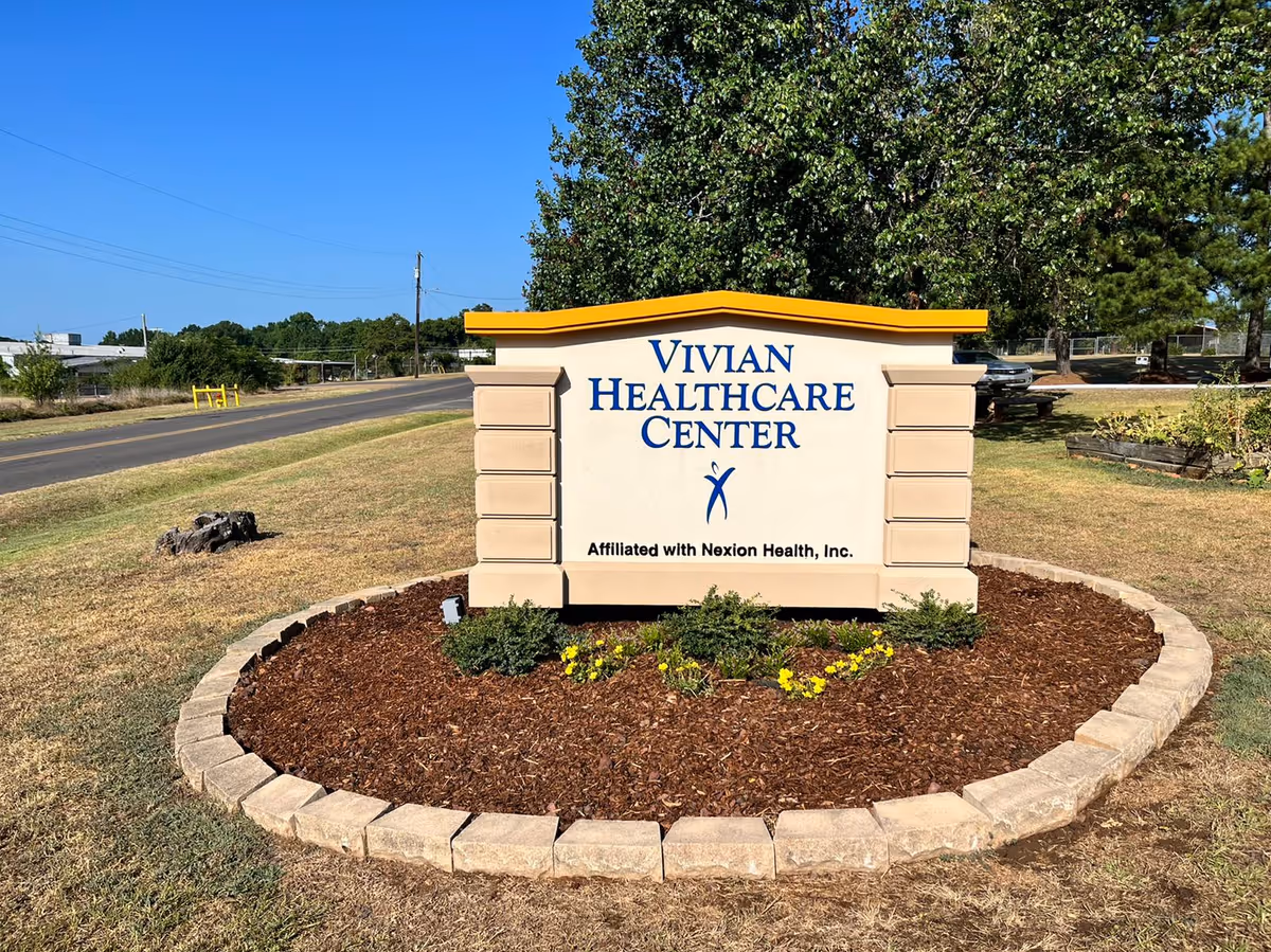 Outdoor view of a sign for Vivian Healthcare Center, surrounded by a circular flower bed with mulch and small plants, located near a road with trees and grass in the background under a clear blue sky.