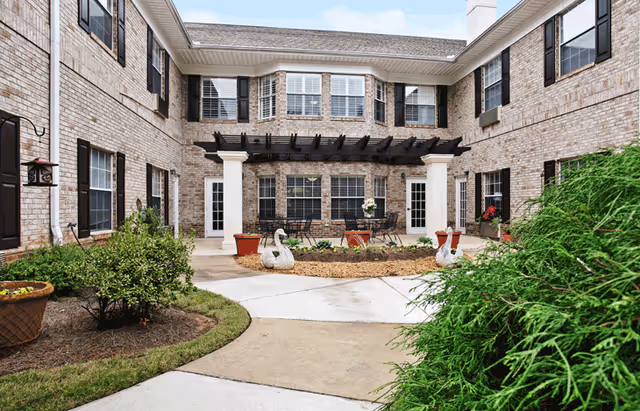 Outdoor courtyard area of a senior living facility with brick walls, multiple windows with black shutters, a pergola over a seating area with tables and chairs, decorative swan statues, potted plants, and landscaped greenery.