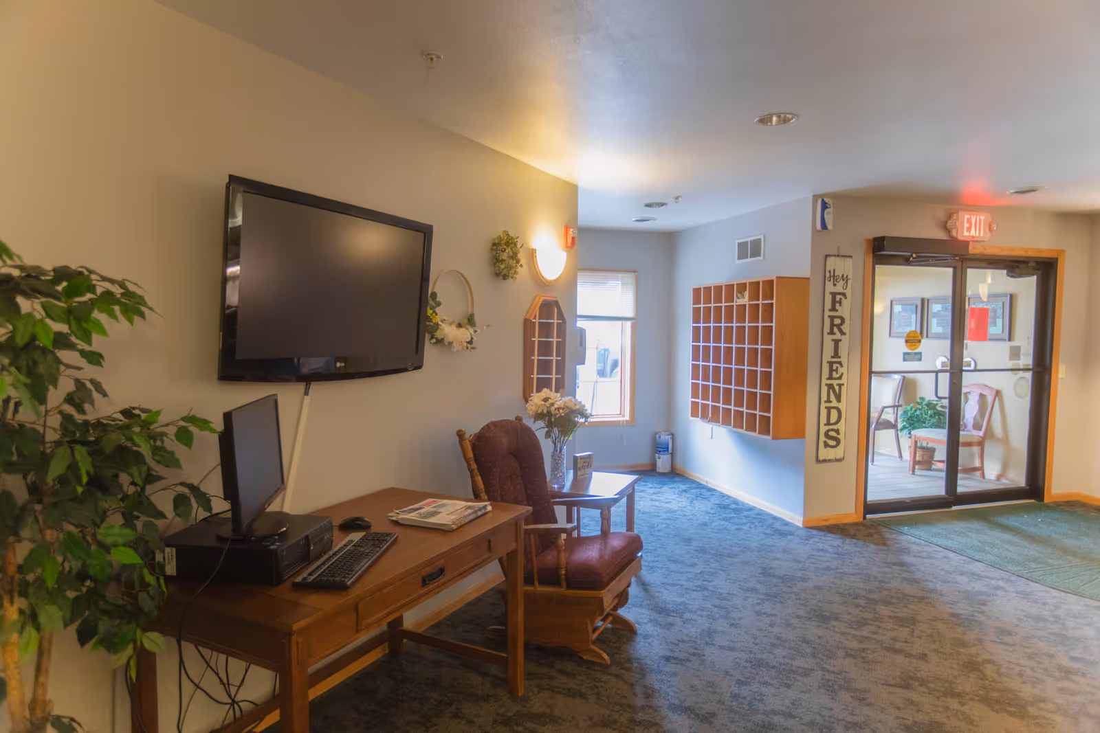 Interior view of a senior living facility lobby area with a wooden desk holding a computer and keyboard, a mounted flat-screen TV on the wall, a cushioned rocking chair, a small table with a flower vase, a plant, a wall-mounted mail organizer, and glass double doors leading outside. A vertical sign next to the door reads 'Hey FRIENDS'.