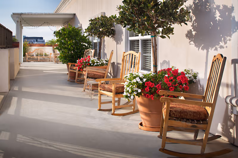 Outdoor patio area with wooden rocking chairs and large potted plants with red and white flowers along a building wall, under a covered pergola with string lights in the background.