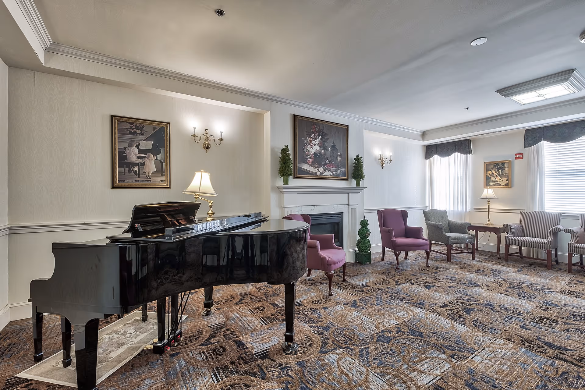 A spacious living room with a grand piano on a patterned carpet. The room features several upholstered chairs in purple and gray, a fireplace with decorative plants on the mantel, framed paintings on the walls, and table lamps providing warm lighting. Large windows with white curtains allow natural light to fill the room.