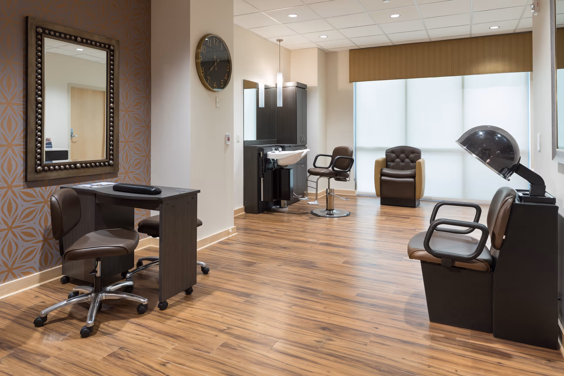 A modern salon area with wooden flooring, featuring a styling chair in front of a sink and mirror, a manicure table with two chairs, a hair dryer station with two chairs, and a comfortable armchair near a large window with blinds.