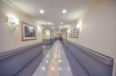 Long, well lit hallway inside Bensonhurst Center for Rehab and Healthcare with framed art and rails.