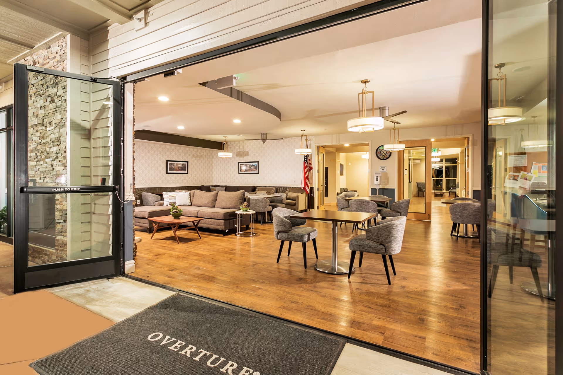 Open entry to a senior living common area with sofas, round tables and chairs on hardwood floors.