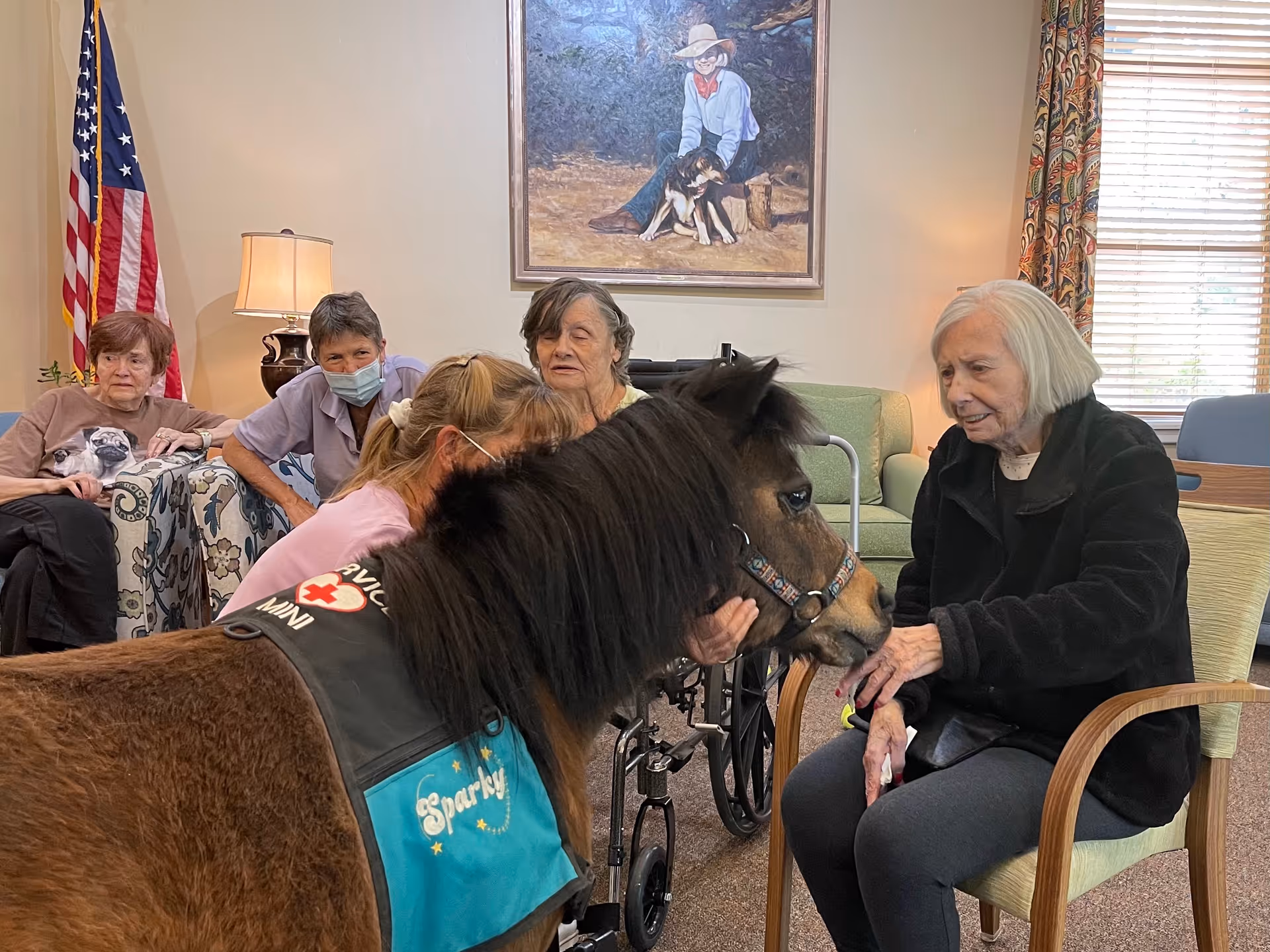 A group of elderly women sitting in a living room area interacting with a small therapy pony named Sparky wearing a vest. The room has an American flag, a lamp, a painting of a person with a dog, and a window with patterned curtains.