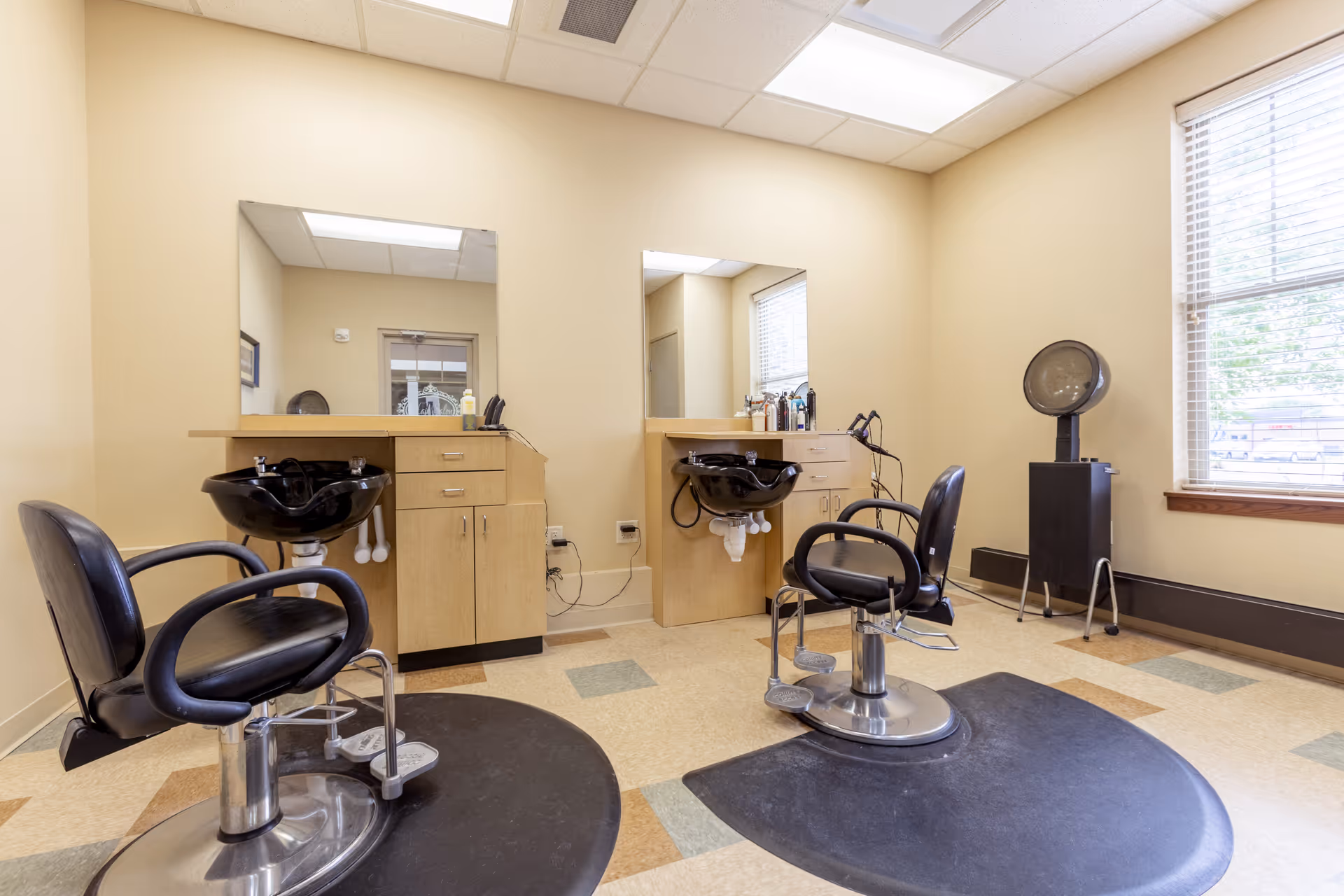 Small salon area with two styling chairs, shampoo sinks, mirrors, a hooded dryer and cabinets by a window.