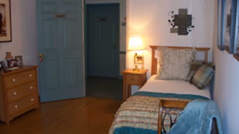 A small bedroom with a single bed against the right wall, covered with a patterned quilt and pillows. A wooden nightstand with a lit lamp is next to the bed. On the left side, there is a wooden dresser with framed photos and a closed blue door. The floor is wooden, and the walls are light-colored with some wall decorations.