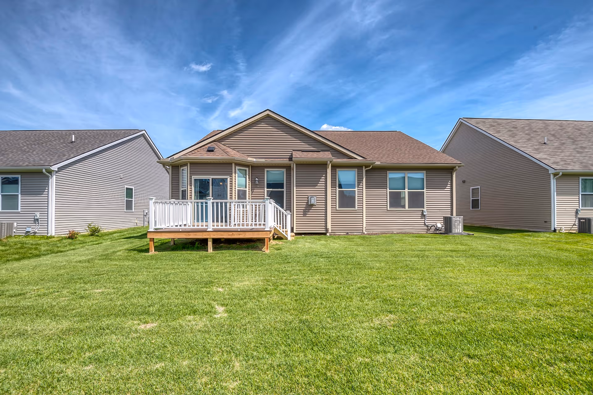 Single-story beige vinyl house with a small raised deck and a large green lawn under a blue sky.