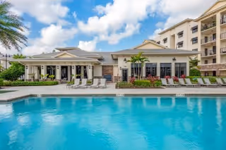 Outdoor swimming pool with clear blue water in front of a single-story building with large windows and a patio area with lounge chairs. Palm trees and a partly cloudy sky are visible in the background.