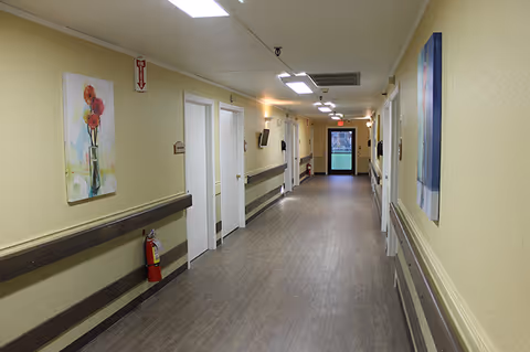 Long beige hallway in a senior living facility with doors, art, and a fire extinguisher.
