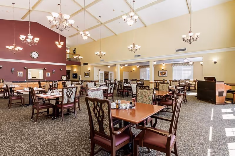 A spacious dining room with multiple wooden tables and upholstered chairs arranged neatly. The room features high ceilings with several hanging chandeliers providing warm lighting. The walls are painted in soft yellow and maroon colors, and there is a clock and framed pictures on the walls. Large windows allow natural light to brighten the space.