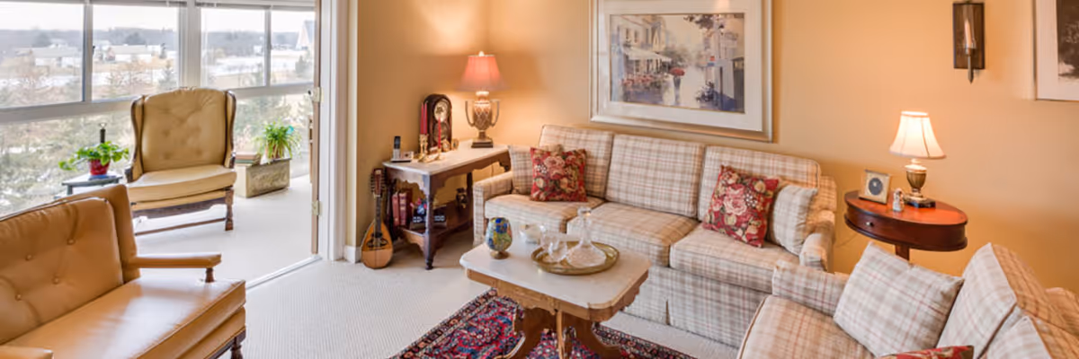 Cozy living room with plaid sofas and armchairs, a central coffee table on a rug, lamps, and large windows opening to a balcony with an outdoor view.