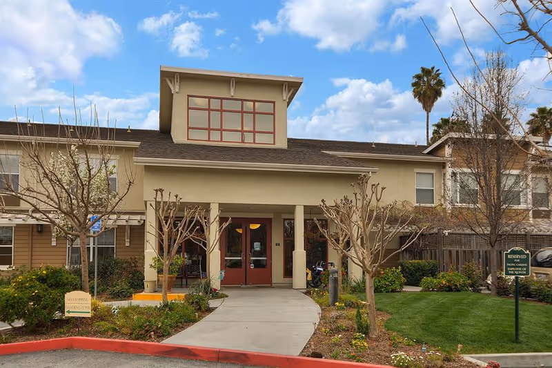 Exterior front view of a senior living facility building with a covered entrance, landscaped garden, and a clear blue sky with some clouds.