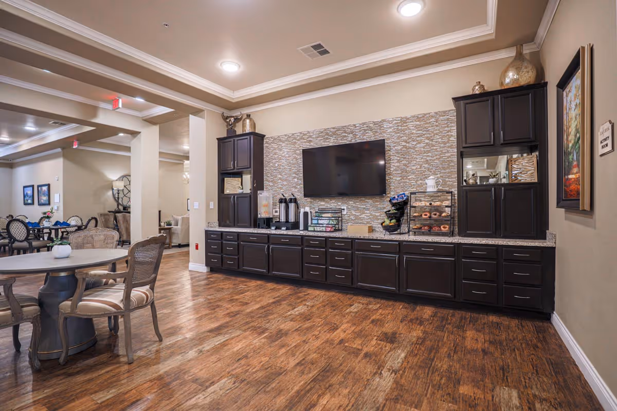 A spacious common area in an assisted living facility featuring a long counter with dark cabinets, a mounted flat-screen TV, coffee and beverage dispensers, and a display case with donuts and fruit. There are round tables with chairs in the foreground and background, and the room has wood flooring and beige walls with decorative molding.