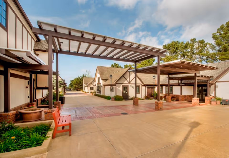 Exterior courtyard and entrance of a senior living community with covered pergolas, paved walkways, benches, and cottage-style buildings under a partly cloudy sky.