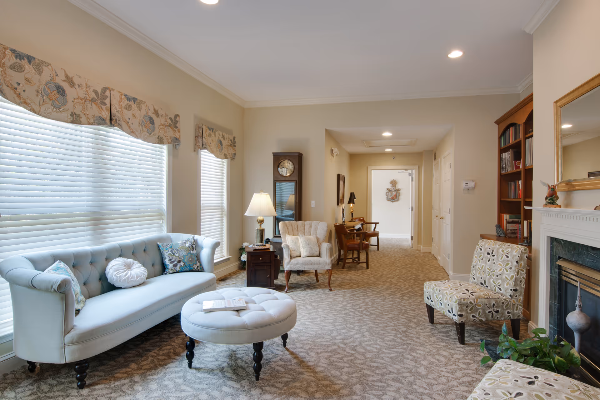 Bright, softly furnished sitting room with tufted sofa, armchairs, ottoman, fireplace and bookcase.