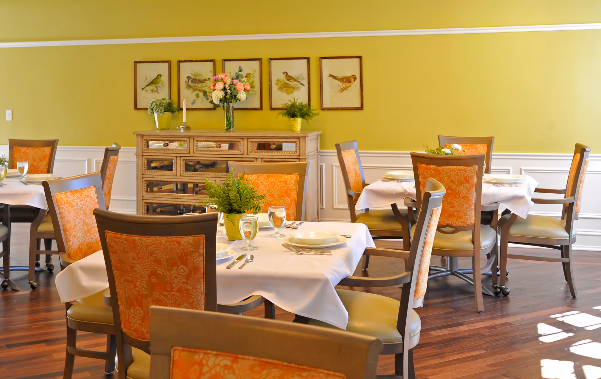 A bright dining room with several tables covered in white tablecloths, each set with plates, glasses, and silverware. The chairs have orange patterned upholstery. A wooden sideboard with decorative plants and a vase of flowers sits against a yellow wall adorned with framed bird illustrations.