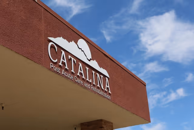 Exterior view of the Catalina Post Acute Care and Rehabilitation building sign mounted on a reddish-brown wall under a blue sky with some clouds.