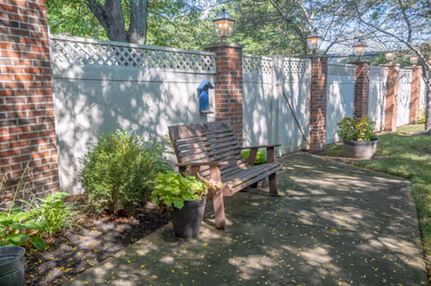 Outdoor patio area with a wooden bench and potted plants along a white fence with brick pillars. Trees and greenery are visible in the background, with sunlight casting shadows on the paved ground.