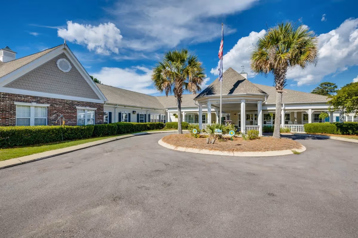 Front exterior view of The Legacy of Lexington facility showing a circular driveway with a landscaped island featuring palm trees and a flagpole, under a partly cloudy blue sky.