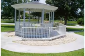 White gazebo with a ramp surrounded by a circular stone border and concrete walkway, set in a grassy outdoor area with trees in the background.