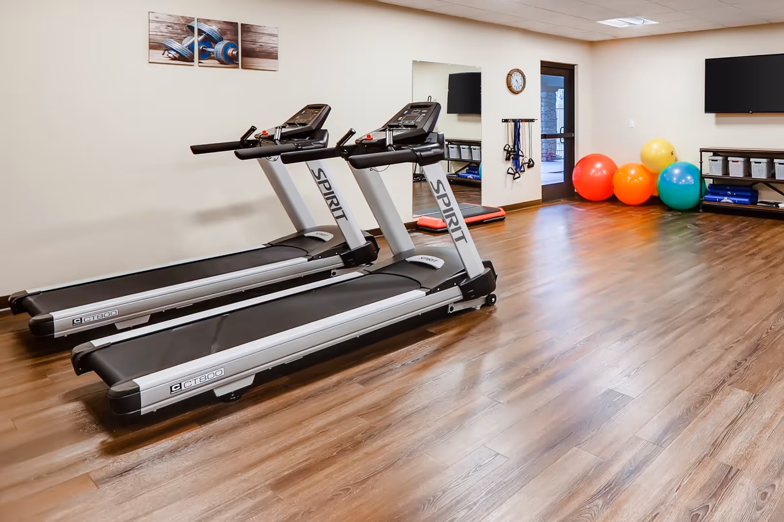 A fitness room with two Spirit brand treadmills on a wooden floor. The room has a wall mirror, a clock, resistance bands hanging on the wall, colorful exercise balls in red, orange, yellow, and blue, and a shelf with storage bins and exercise mats. A door with a window leads outside.