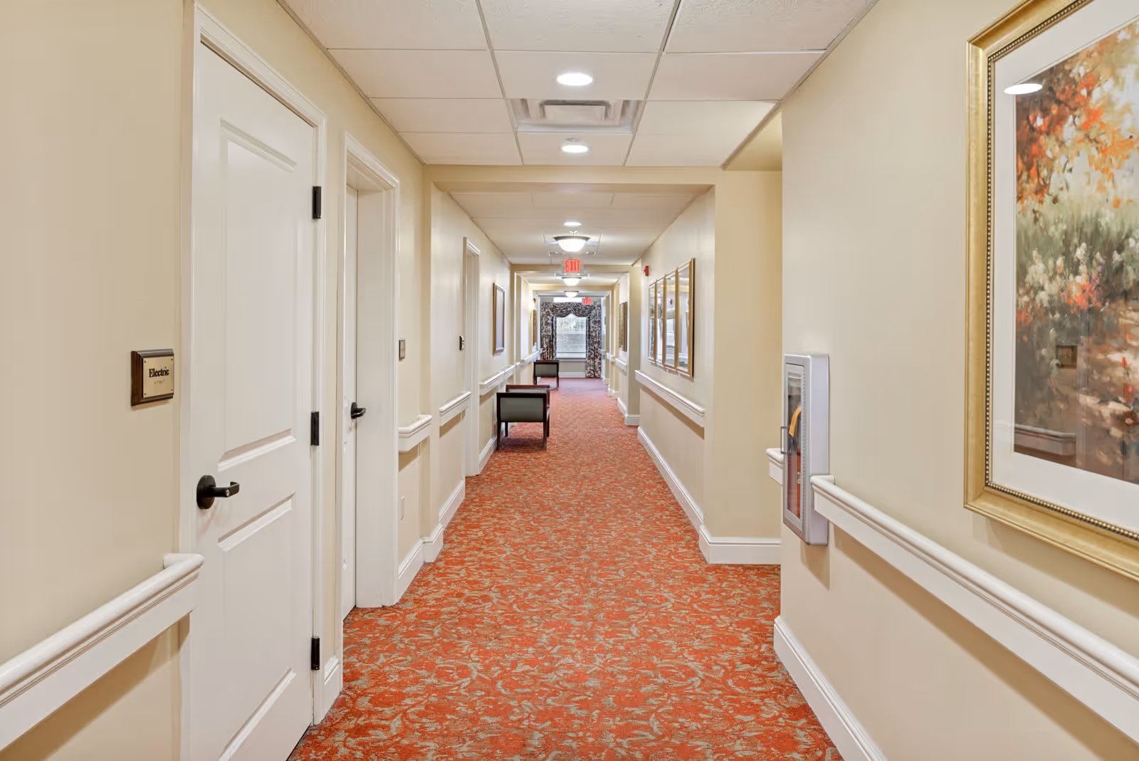 A long, well-lit hallway in a senior living facility with beige walls, white doors, and a red patterned carpet. The hallway features handrails on both sides, framed artwork on the walls, and a few chairs placed along the corridor. Ceiling lights illuminate the space evenly.